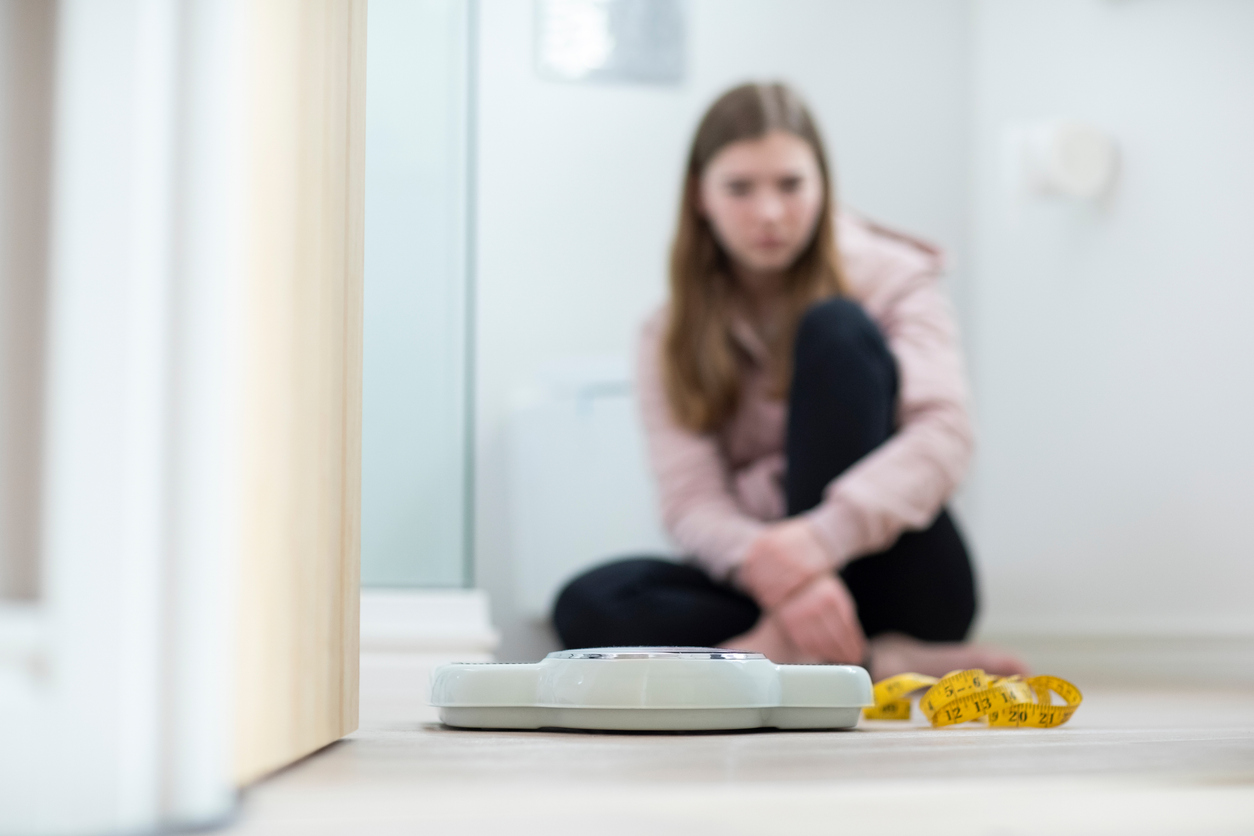 Unhappy Teenage Girl Sitting In Bathroom Looking At Scales And Tape Measure.
